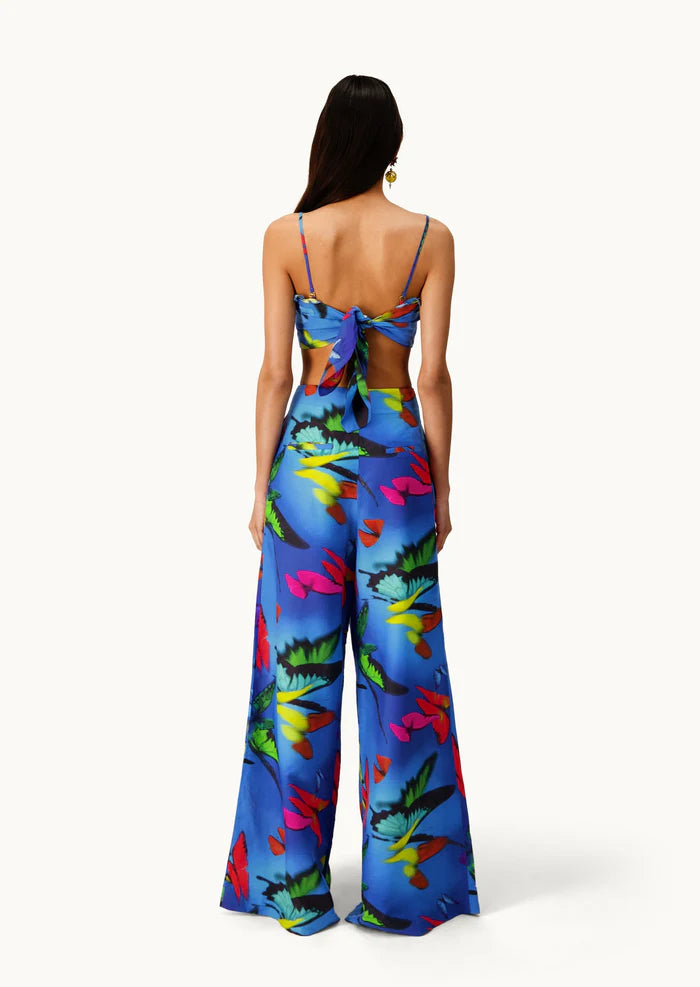 A woman faces away, wearing the Alma Set—a blue floral jumpsuit with wide-leg pants and an open back finished with a statement bow. Her long, straight hair pairs with a single hoop earring, showcasing the set’s versatile styling that can be effortlessly elevated for any occasion with adaptable accessories.