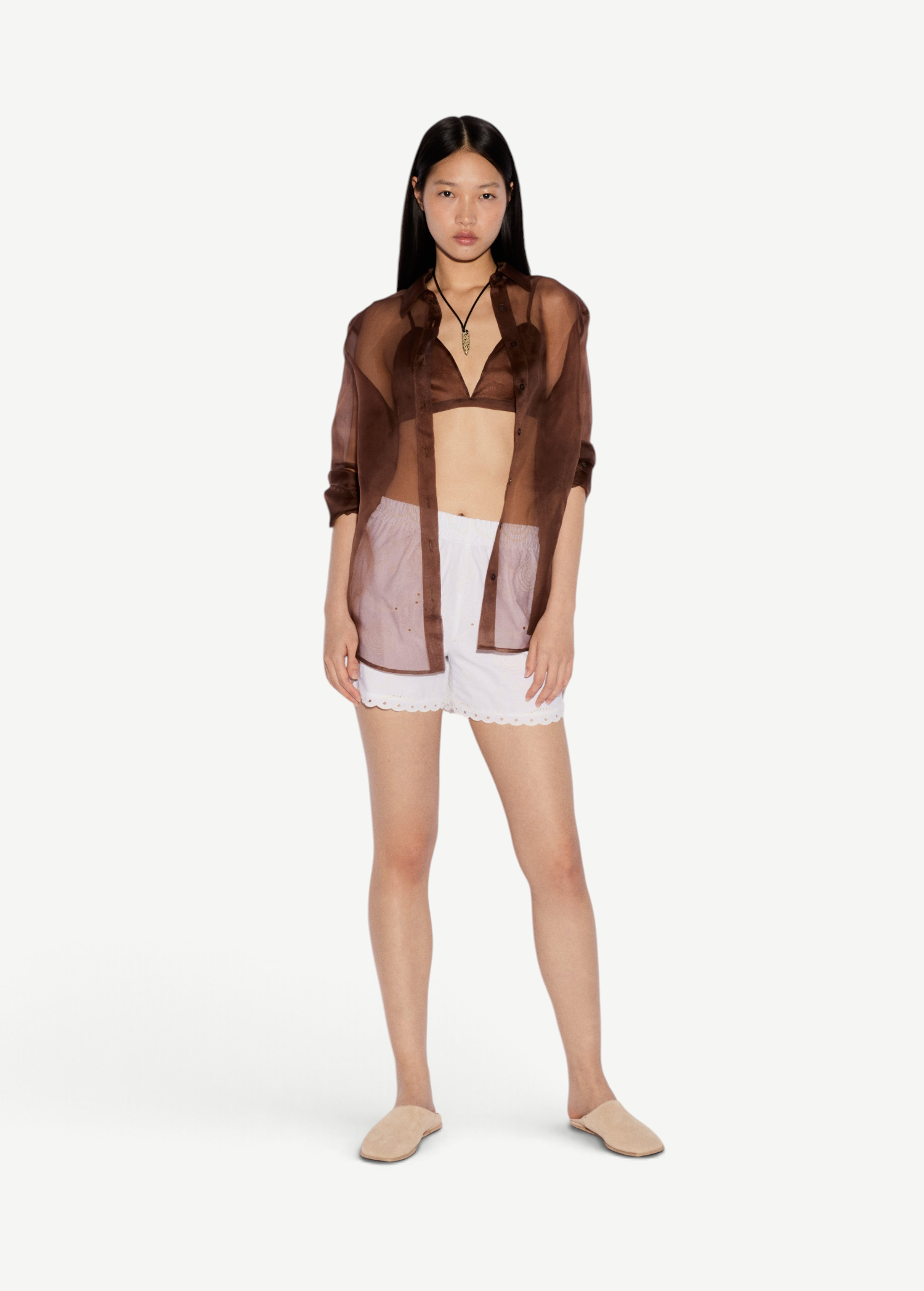 Model wearing KABE SHOP INC Kira white shorts with scalloped hem, paired with a brown sheer shirt and beige mules.