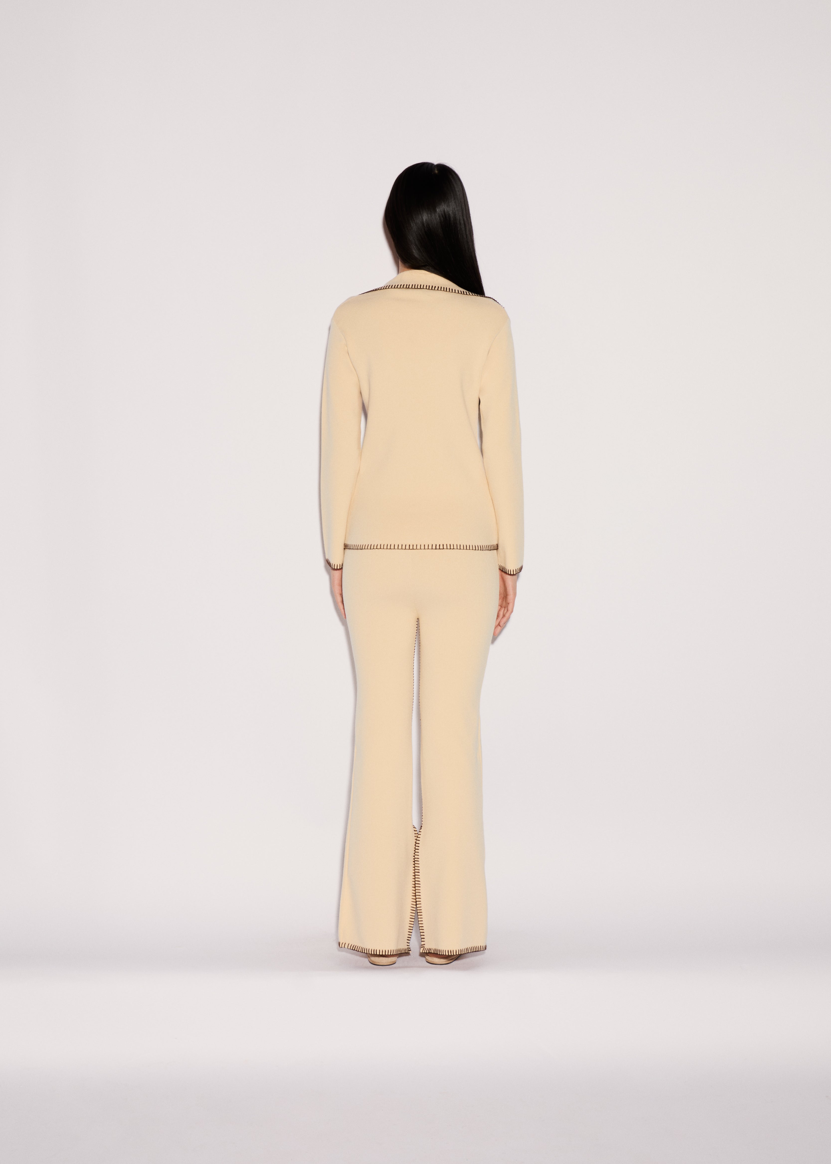 Back view of a person in a relaxed silhouette wearing a cream-colored Iris Top and matching flared trousers, both finished with decorative brown whipstitching along the hems and edges.