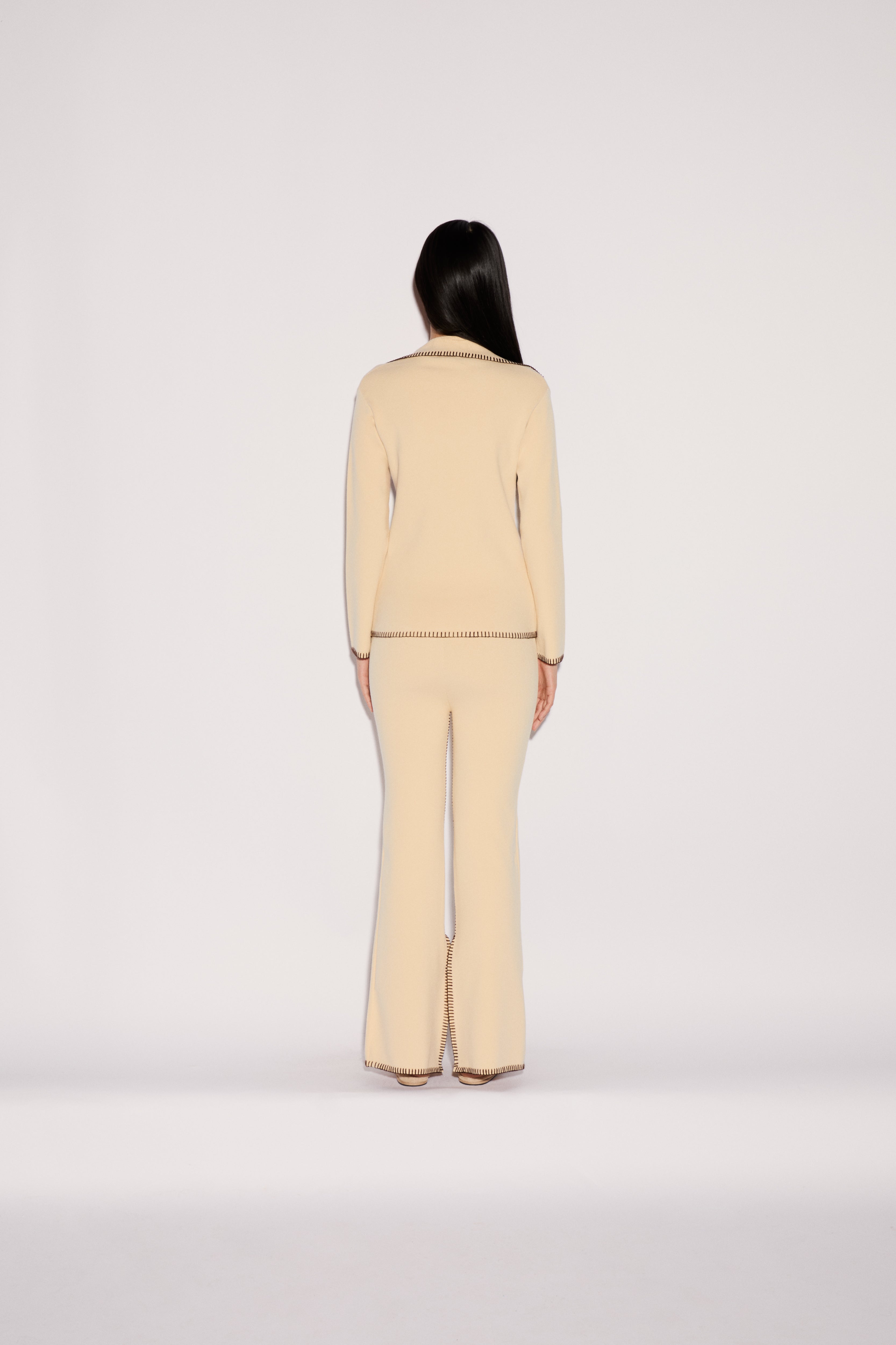 Back view of a person in a relaxed silhouette wearing a cream-colored Iris Top and matching flared trousers, both finished with decorative brown whipstitching along the hems and edges.