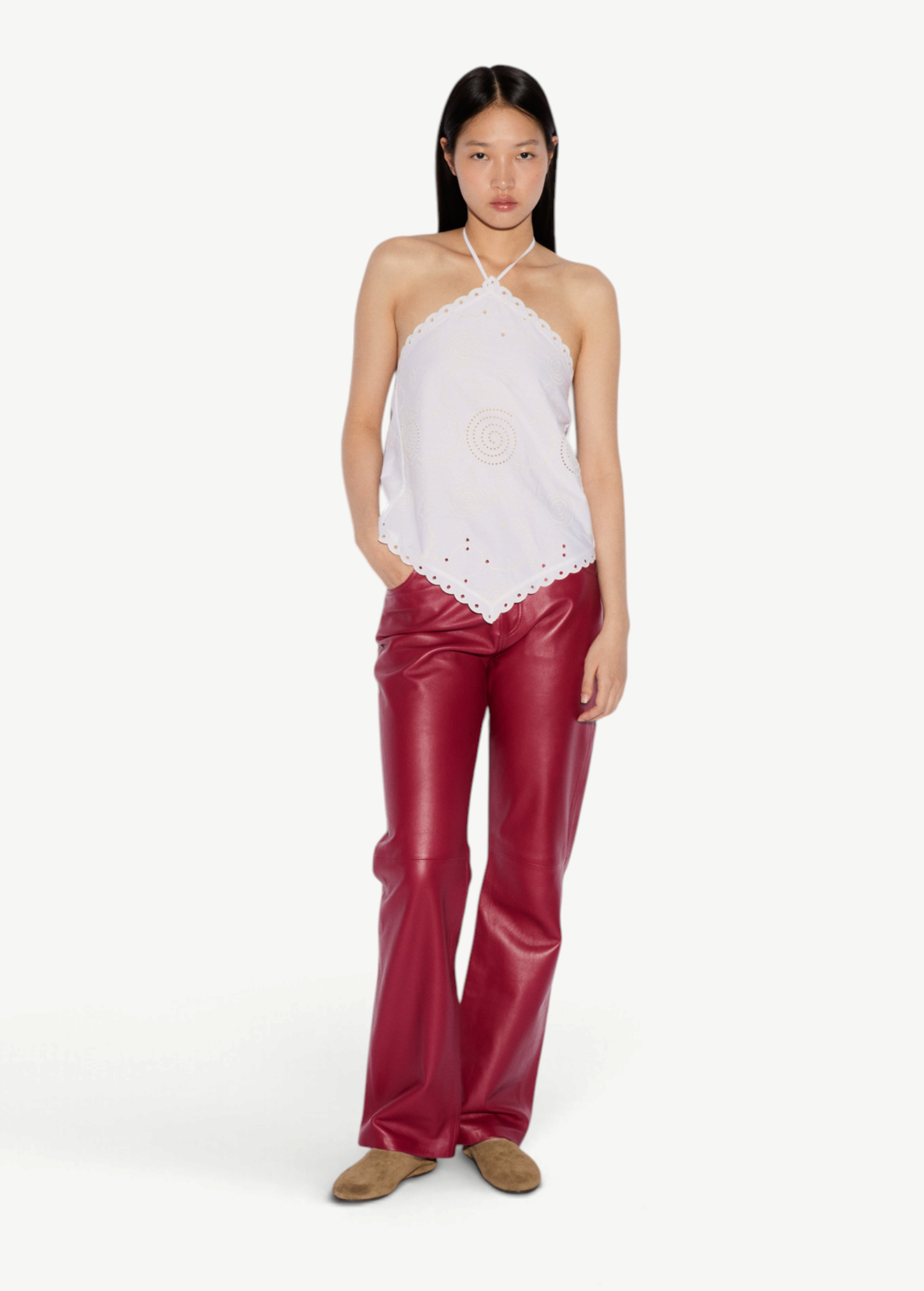 A person wears an elegant, white Cleo Top with a halter-neck and eyelet embroidery, paired with red trousers and tan shoes.