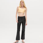 A woman with long blonde hair wears a beige crop top, the Xena Pants in black leather, and black sandals, standing against a plain white background.