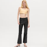 A woman with long blonde hair wears a beige crop top, the Xena Pants in black leather, and black sandals, standing against a plain white background.