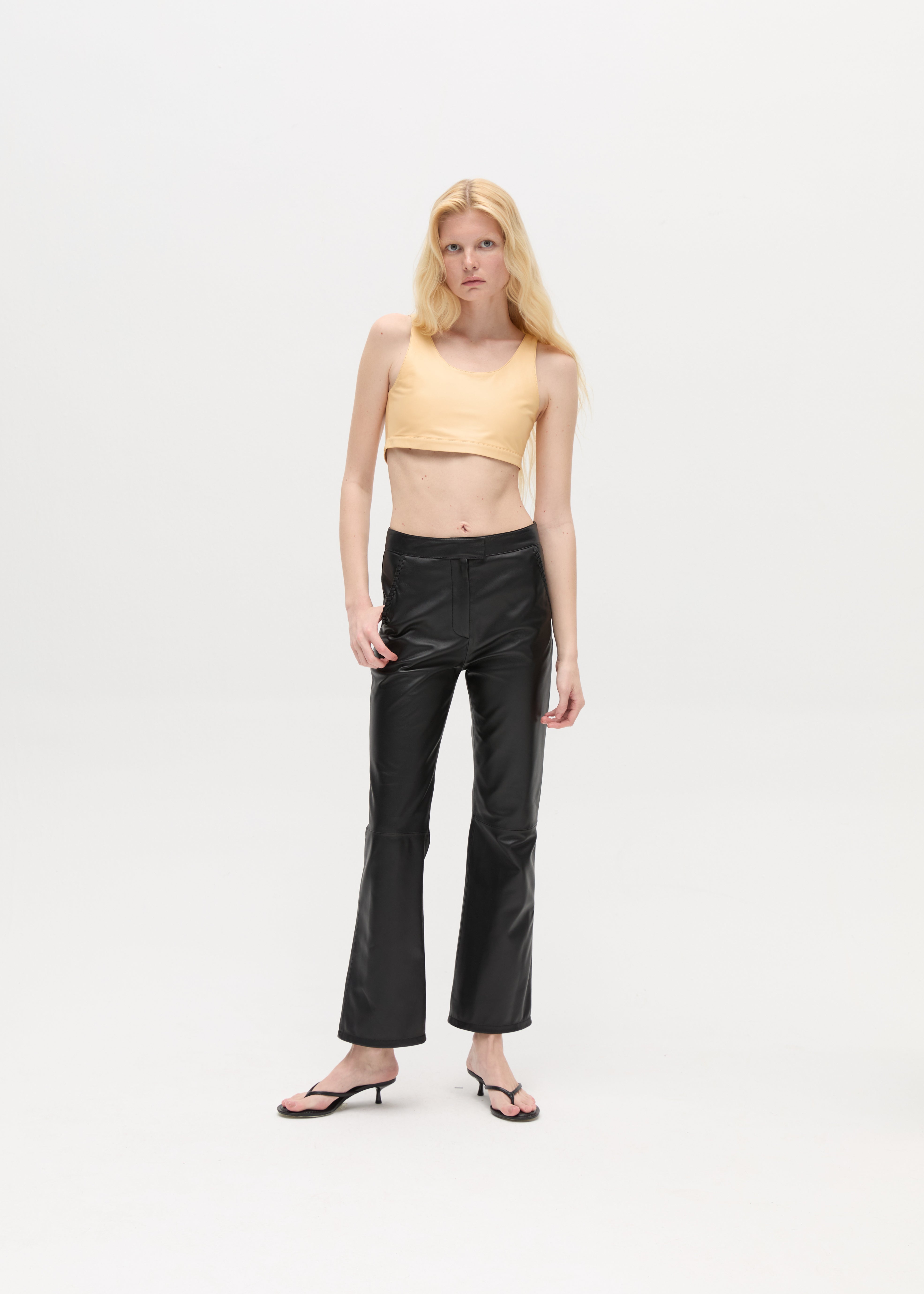 A woman with long blonde hair wears a beige crop top, the Xena Pants in black leather, and black sandals, standing against a plain white background.