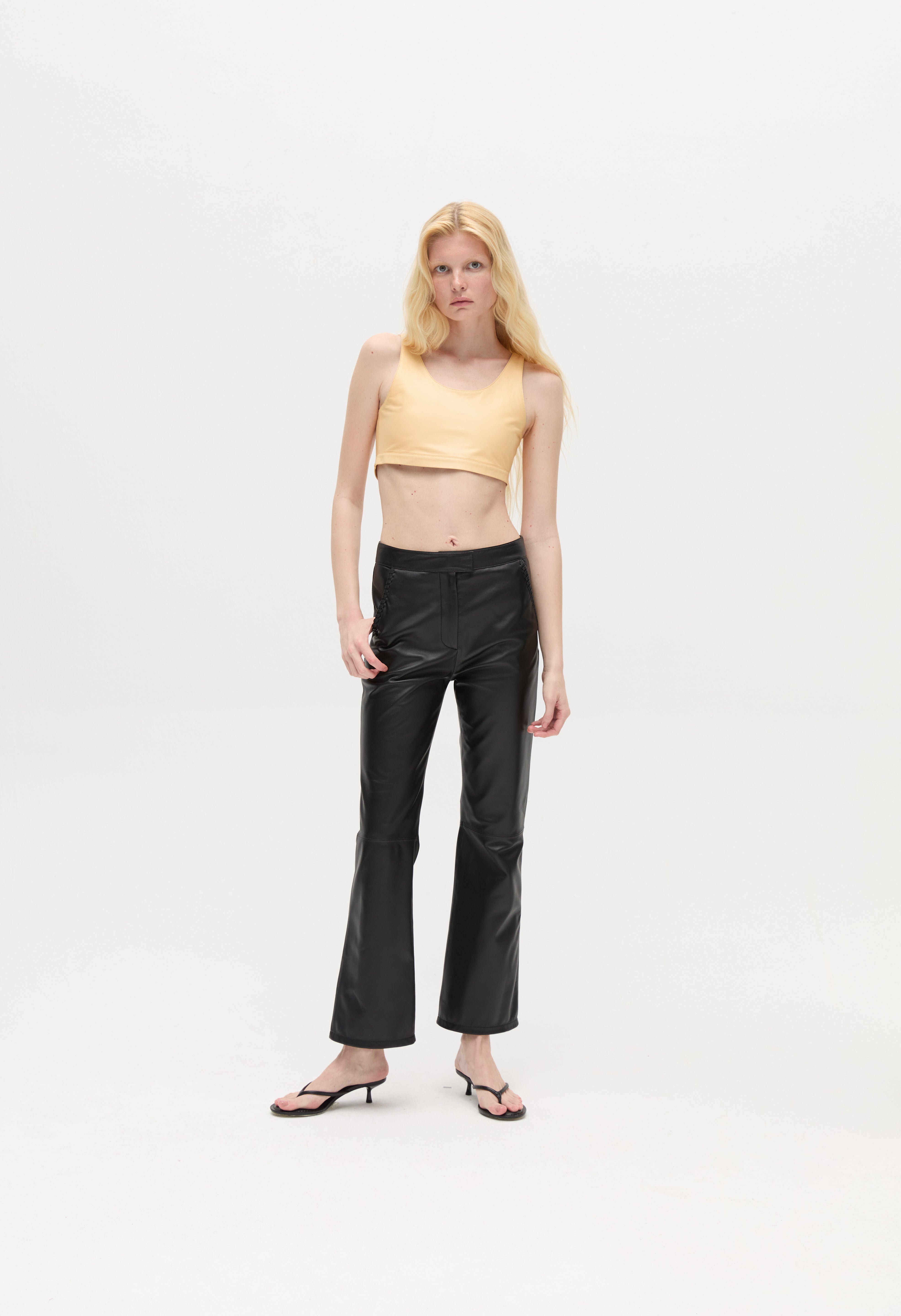 A woman with long blonde hair wears a beige crop top, the Xena Pants in black leather, and black sandals, standing against a plain white background.