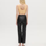 A person with long blonde hair is standing facing away, wearing a peach sleeveless top, the Xena Pants in black leather, and black heeled sandals against a plain white background.
