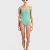 A woman stands barefoot against a plain white background, wearing the Margot One Piece Swimwear in light green. The luxury swimsuit is crafted from premium Embodee fabric and features wide straps with a square neckline. She faces forward with a neutral expression, her arms relaxed at her sides.