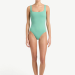 A woman stands barefoot against a plain white background, wearing the Margot One Piece Swimwear in light green. The luxury swimsuit is crafted from premium Embodee fabric and features wide straps with a square neckline. She faces forward with a neutral expression, her arms relaxed at her sides.