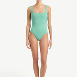 A woman stands barefoot against a plain white background, wearing the Margot One Piece Swimwear in light green. The luxury swimsuit is crafted from premium Embodee fabric and features wide straps with a square neckline. She faces forward with a neutral expression, her arms relaxed at her sides.