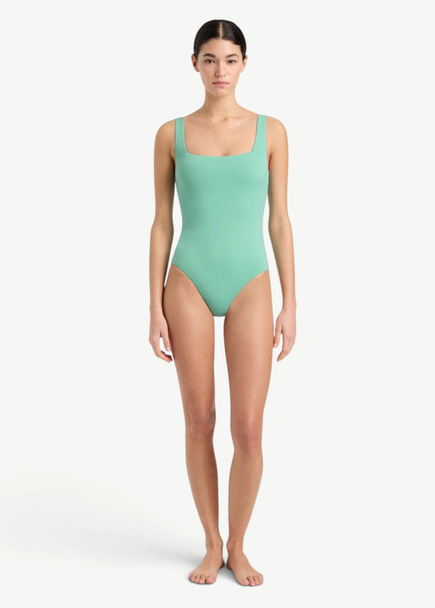 A woman stands barefoot against a plain white background, wearing the Margot One Piece Swimwear in light green. The luxury swimsuit is crafted from premium Embodee fabric and features wide straps with a square neckline. She faces forward with a neutral expression, her arms relaxed at her sides.