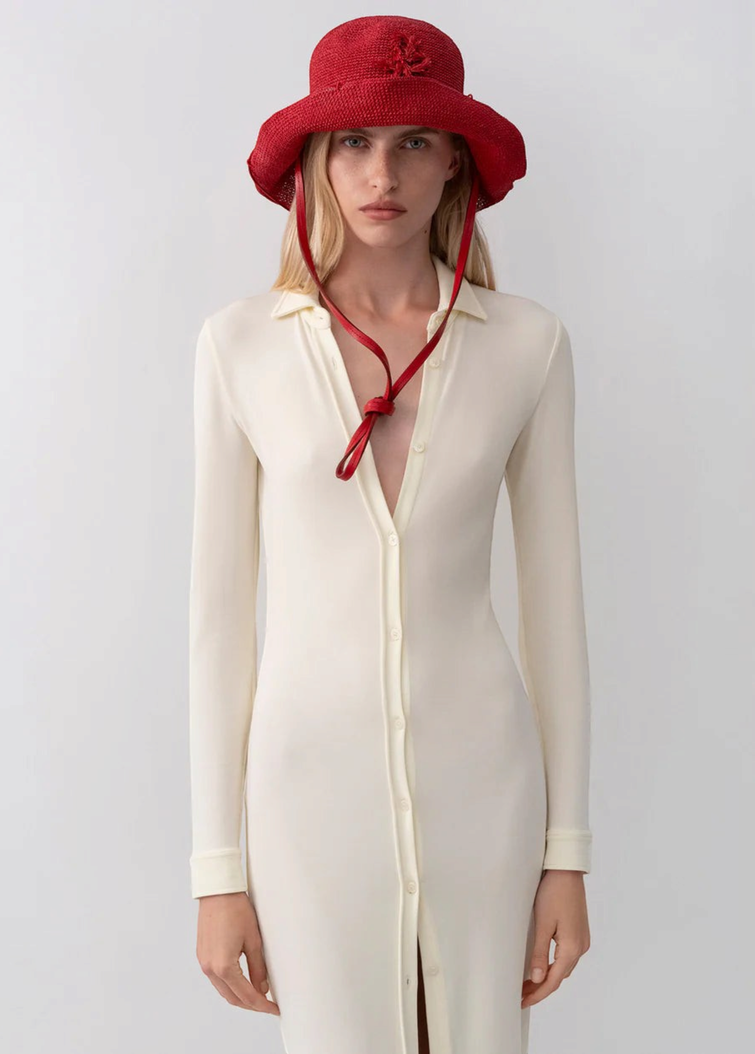 Model wearing a red KABE SHOP INC straw hat bag with a long chin strap, styled with a cream button-down midi dress.