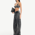 A woman stands confidently against a plain white background, wearing Acid Wash Boyfriend Jeans paired with a dark bralette and white open-toe shoes. She holds a black clutch in one hand, her long straight hair framing her look.