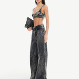 A woman stands confidently against a plain white background, wearing Acid Wash Boyfriend Jeans paired with a dark bralette and white open-toe shoes. She holds a black clutch in one hand, her long straight hair framing her look.