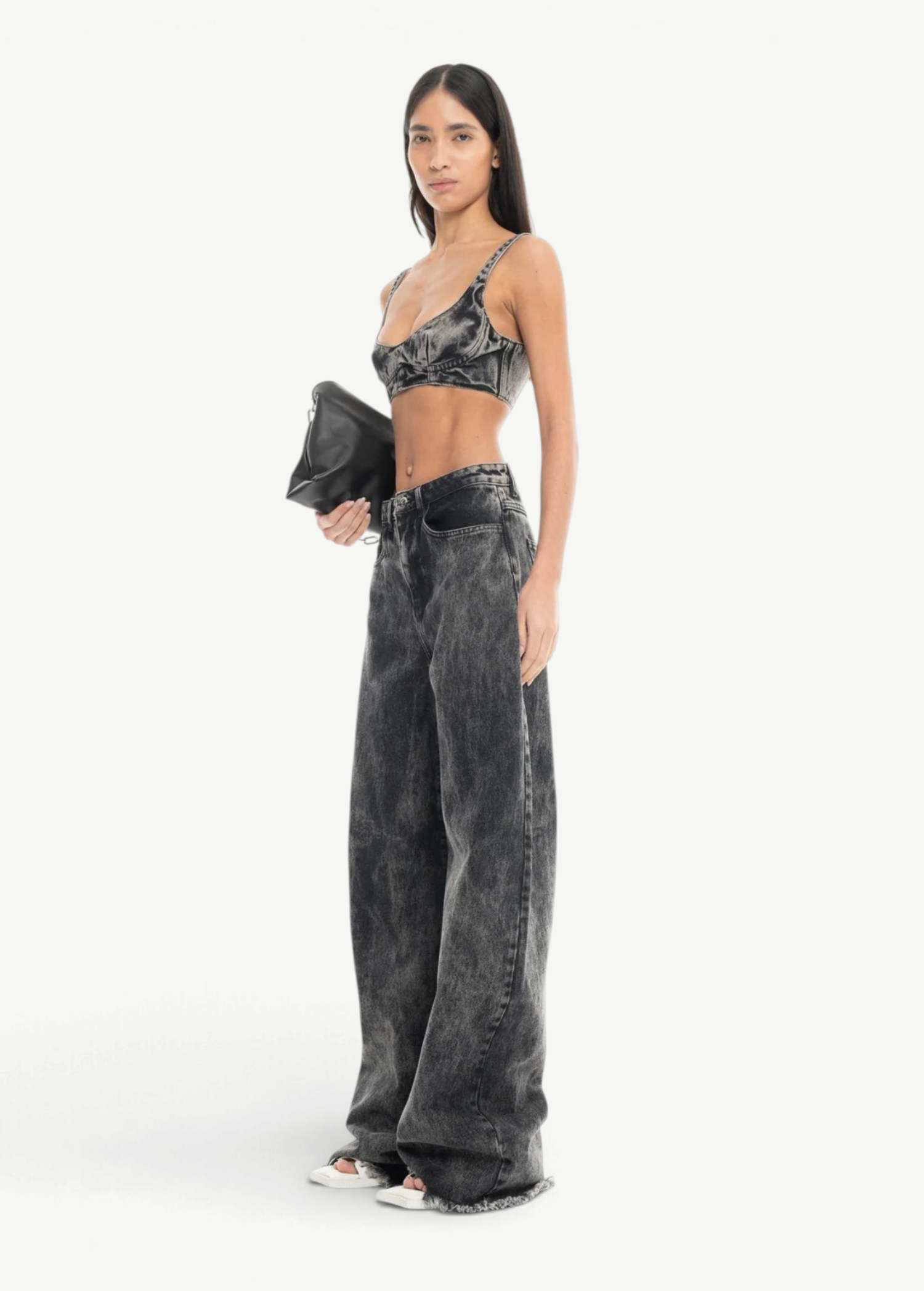 A woman stands confidently against a plain white background, wearing Acid Wash Boyfriend Jeans paired with a dark bralette and white open-toe shoes. She holds a black clutch in one hand, her long straight hair framing her look.