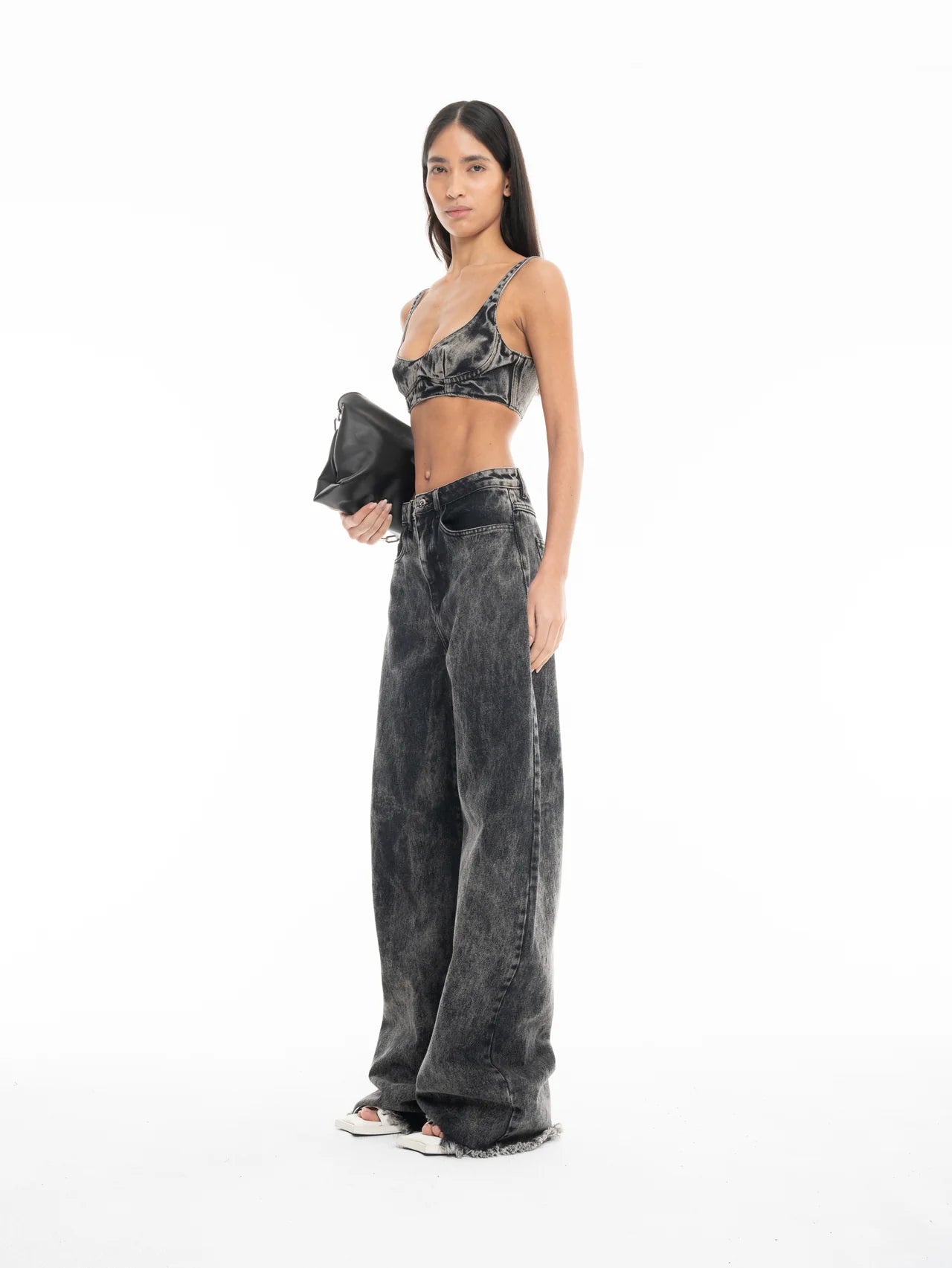 A woman stands confidently against a plain white background, wearing Acid Wash Boyfriend Jeans paired with a dark bralette and white open-toe shoes. She holds a black clutch in one hand, her long straight hair framing her look.