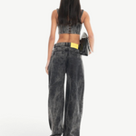 A person with long dark hair, wearing a sleeveless crop top and the Acid Wash Boyfriend Jeans, stands facing away while holding a black bag against a white background.