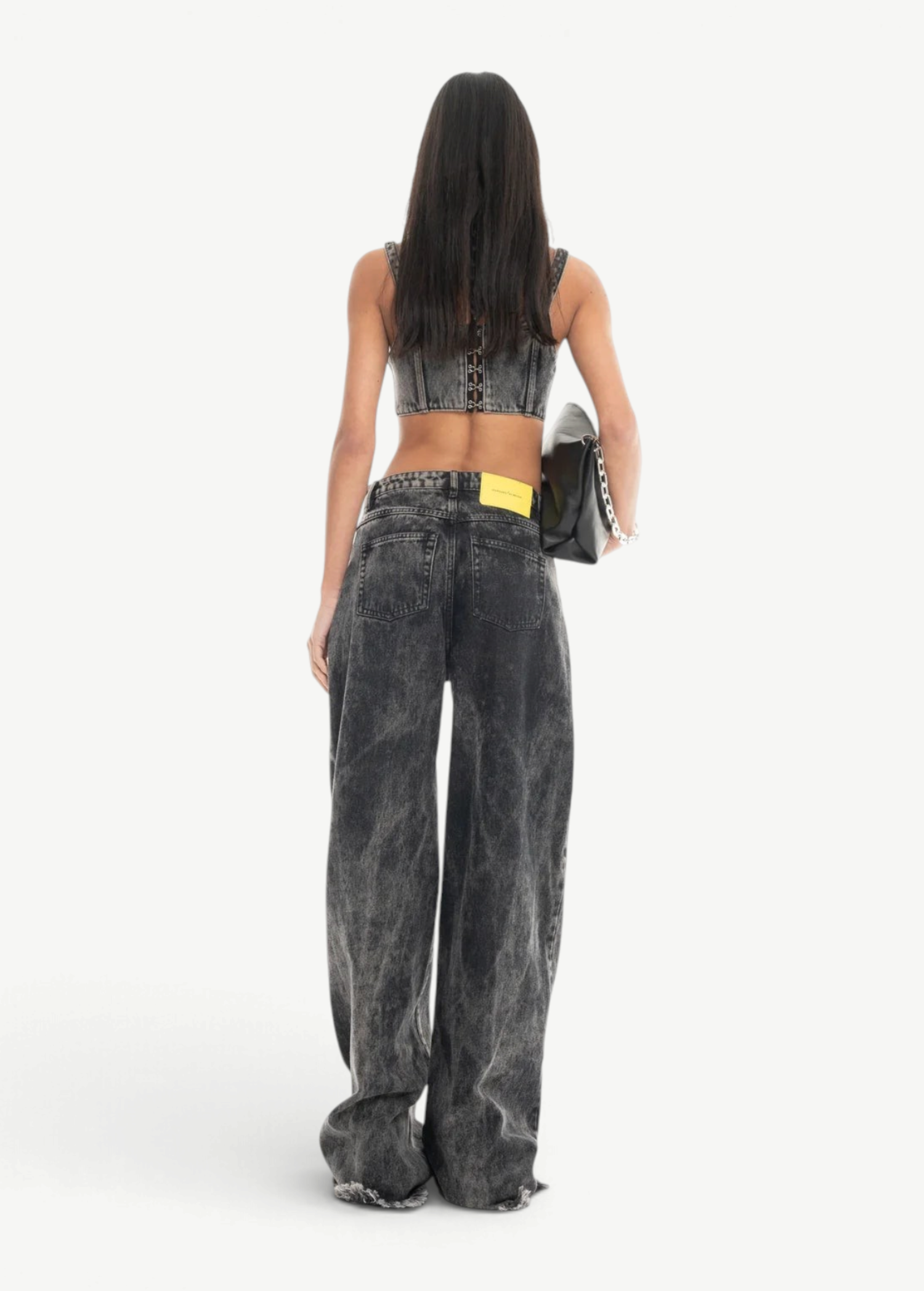 A person with long dark hair, wearing a sleeveless crop top and the Acid Wash Boyfriend Jeans, stands facing away while holding a black bag against a white background.