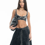 A woman with long hair stands against a white background, wearing the Denim Bra Corset and matching skirt, and holding a black bag under her arm.