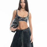 A woman with long hair stands against a white background, wearing the Denim Bra Corset and matching skirt, and holding a black bag under her arm.