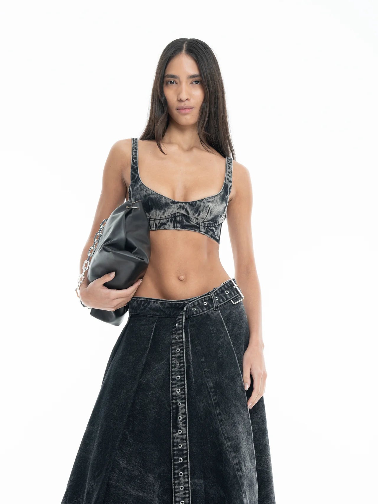 A woman with long hair stands against a white background, wearing the Denim Bra Corset and matching skirt, and holding a black bag under her arm.