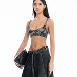 A woman wearing a black acid-wash Denim Bra Corset and matching skirt holds a black clutch bag, posing against a white background.