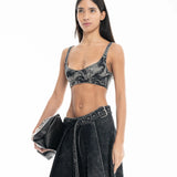 A woman wearing a black acid-wash Denim Bra Corset and matching skirt holds a black clutch bag, posing against a white background.