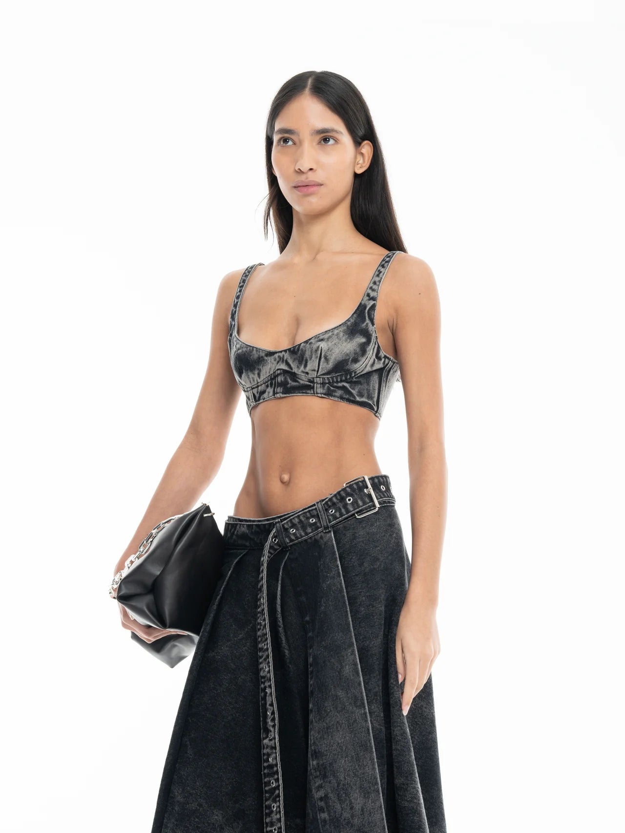 A woman wearing a black acid-wash Denim Bra Corset and matching skirt holds a black clutch bag, posing against a white background.