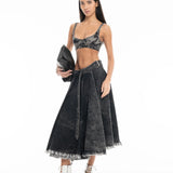A woman wearing a black Denim Bra Corset and coordinating high-waisted, flared skirt with frayed hems holds a black clutch bag and pairs the outfit with white lace-up heels, posing against a plain white background.