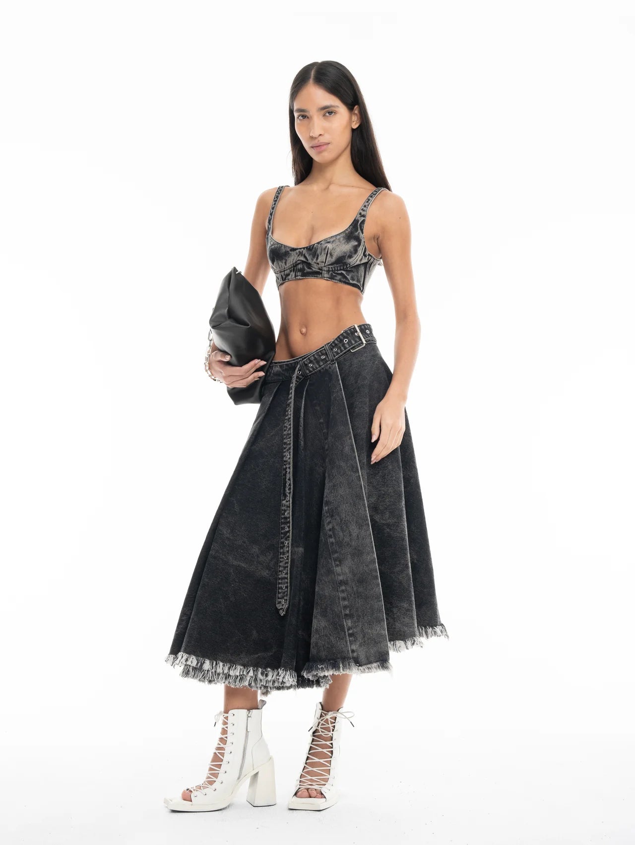 A woman wearing a black Denim Bra Corset and coordinating high-waisted, flared skirt with frayed hems holds a black clutch bag and pairs the outfit with white lace-up heels, posing against a plain white background.