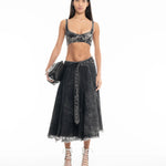 A woman stands against a white background wearing a black denim bralette, the Midi Kilt Skirt with Buckle Fastening in black acid wash with a frayed hem, white lace-up heels, and holding a black handbag.