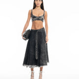 A woman stands against a white background wearing a black denim bralette, the Midi Kilt Skirt with Buckle Fastening in black acid wash with a frayed hem, white lace-up heels, and holding a black handbag.