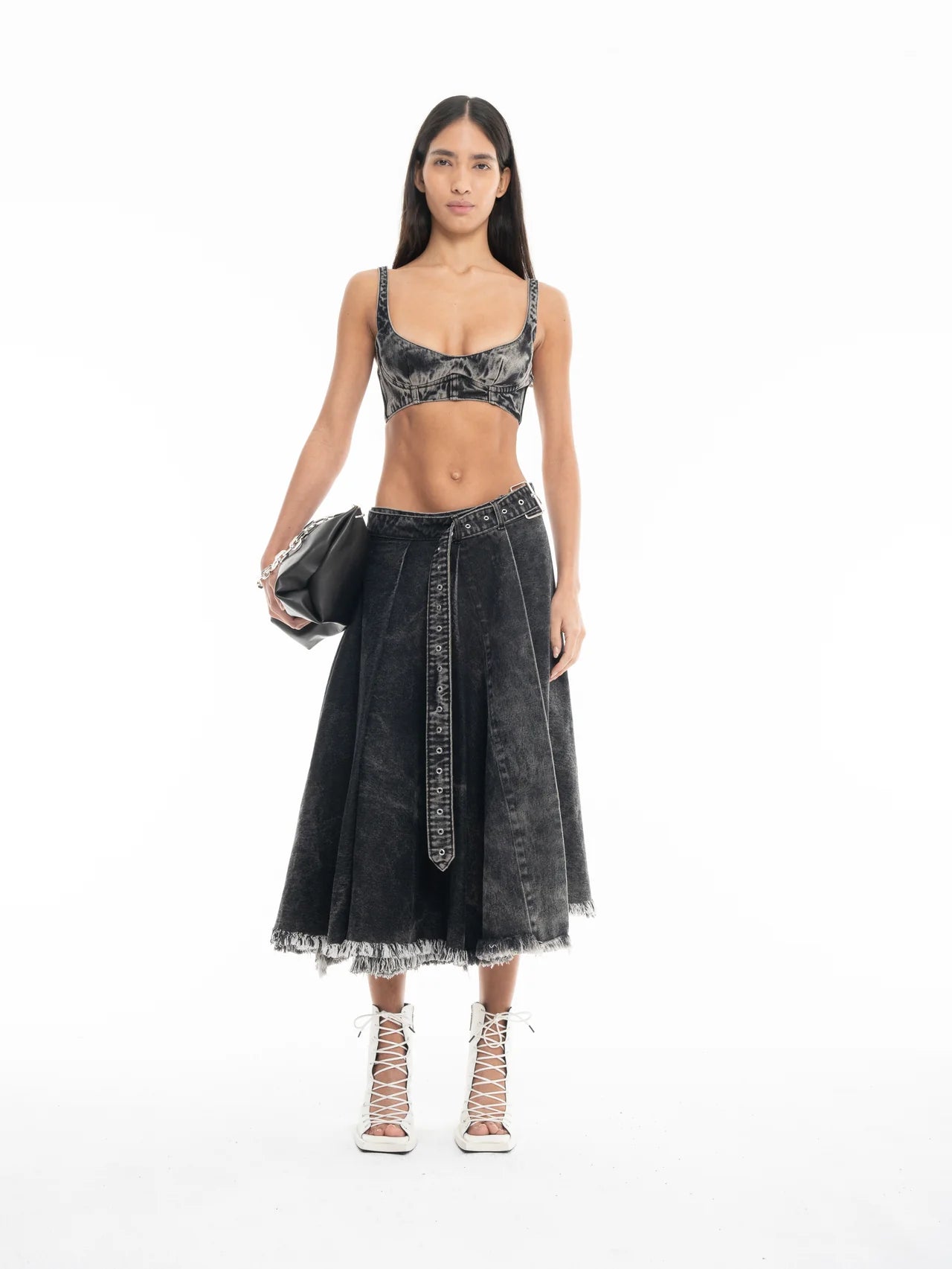 A woman stands against a white background wearing a black denim bralette, the Midi Kilt Skirt with Buckle Fastening in black acid wash with a frayed hem, white lace-up heels, and holding a black handbag.