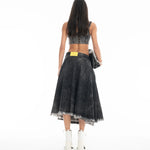 A woman with long dark hair stands facing away, wearing a black sleeveless top, the Midi Kilt Skirt with Buckle Fastening in black acid wash featuring a yellow patch, white ankle boots, and holding a black bag against a white background.