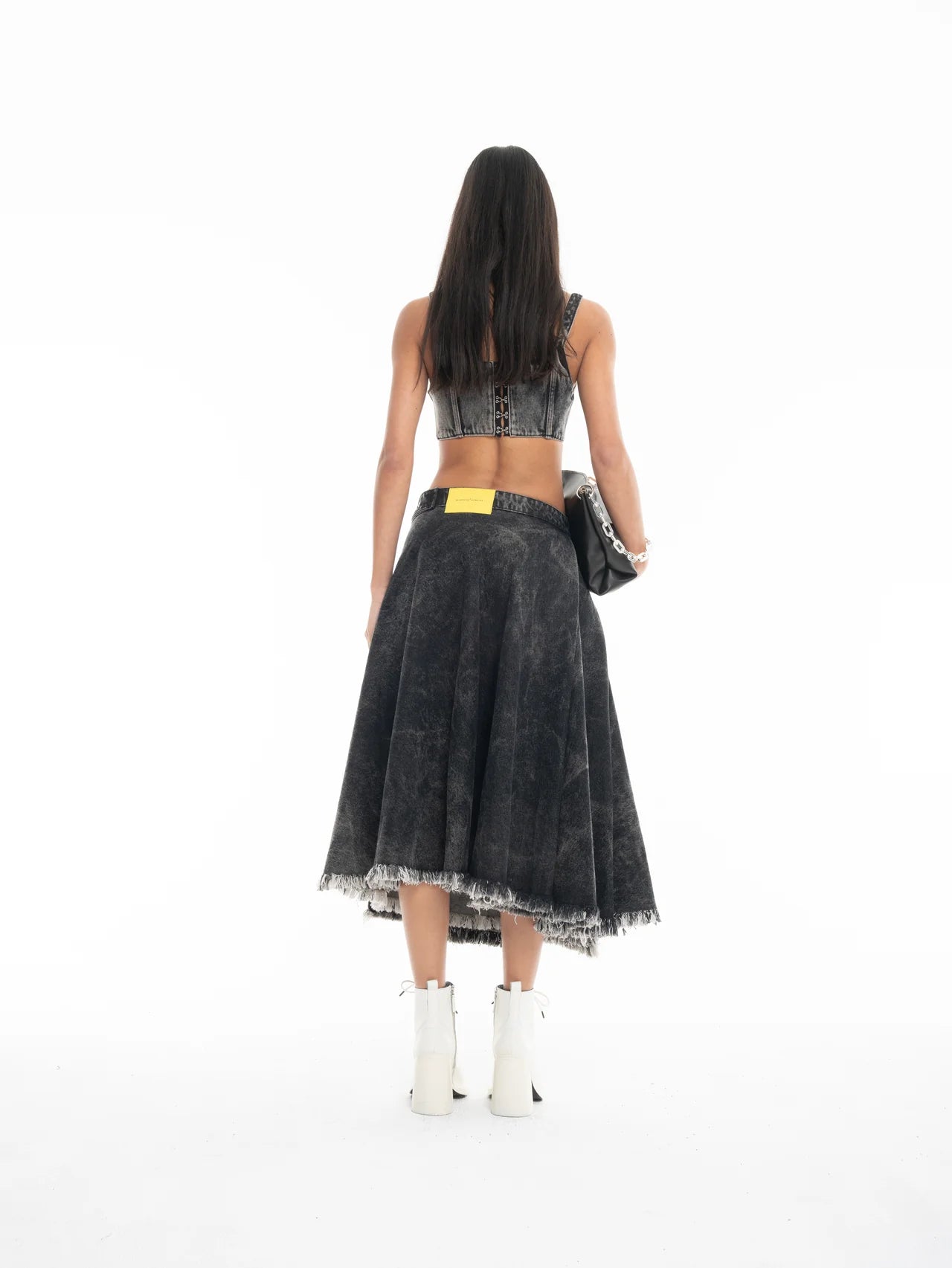 A woman with long dark hair stands facing away, wearing a black sleeveless top, the Midi Kilt Skirt with Buckle Fastening in black acid wash featuring a yellow patch, white ankle boots, and holding a black bag against a white background.