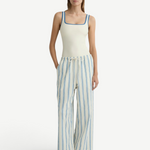 A model wearing a white tank top with blue trim and lightweight, white Finn Pant wide-leg drawstring pants with vertical blue stripes.