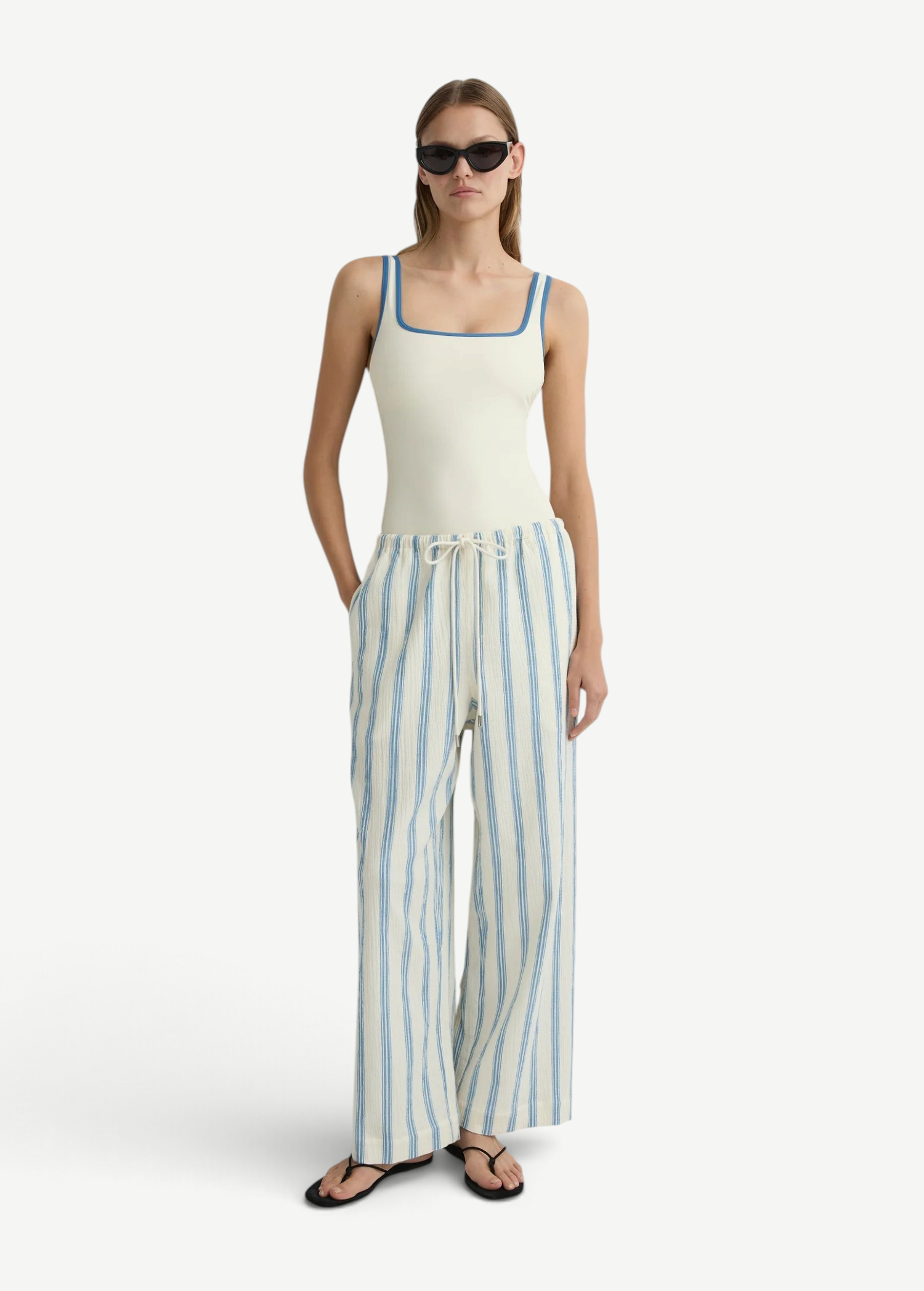 A model wearing a white tank top with blue trim and lightweight, white Finn Pant wide-leg drawstring pants with vertical blue stripes.