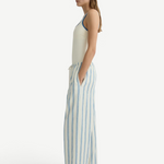 A side view of a person wearing a cream-colored tank top and Finn Pant, which are lightweight, loose-fitting cream pants featuring vertical blue stripes and a drawstring waist.