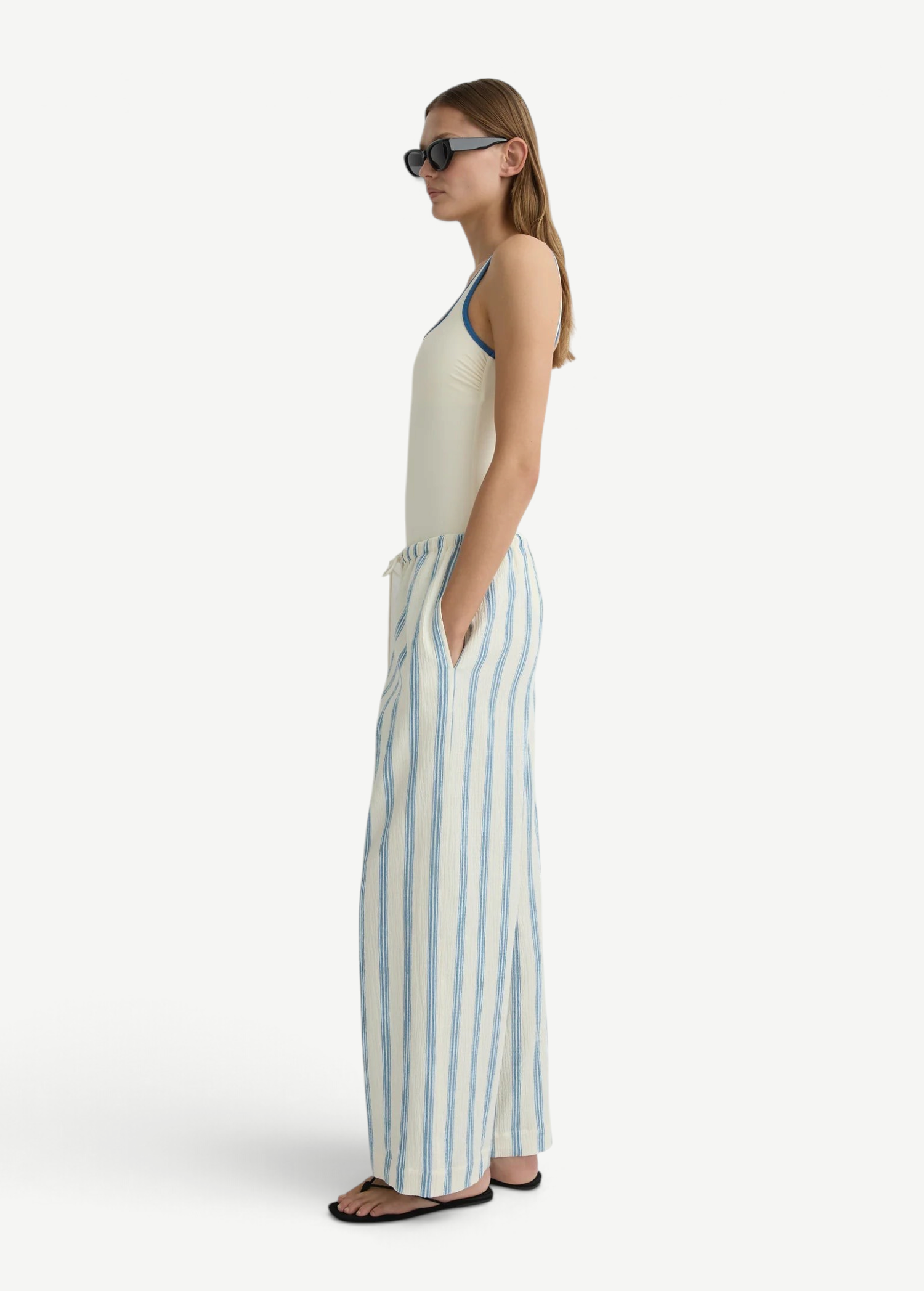 A side view of a person wearing a cream-colored tank top and Finn Pant, which are lightweight, loose-fitting cream pants featuring vertical blue stripes and a drawstring waist.