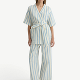 A woman wearing cream Finn Pant drawstring pants with a matching short-sleeved blue-striped wrap top and black flip-flops.