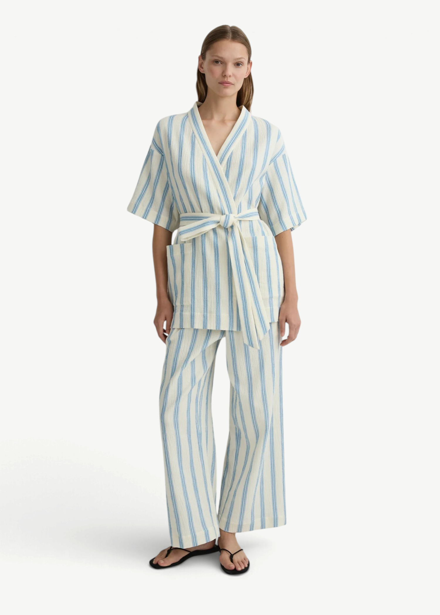 A woman wearing cream Finn Pant drawstring pants with a matching short-sleeved blue-striped wrap top and black flip-flops.