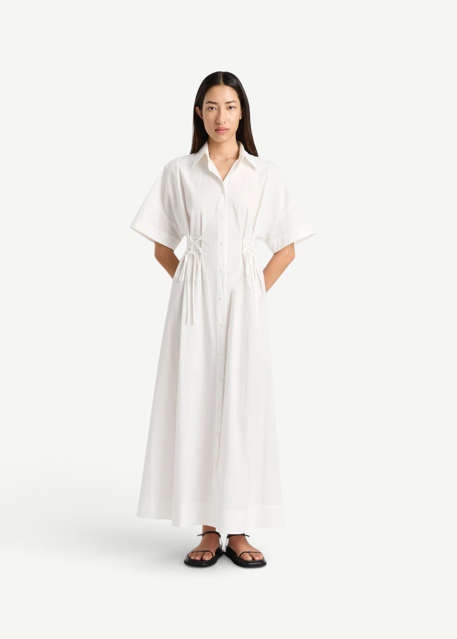 Model wearing a white KABE SHOP INC Kyoto lace-up shirt dress with short sleeves and a 7/8 length hem.