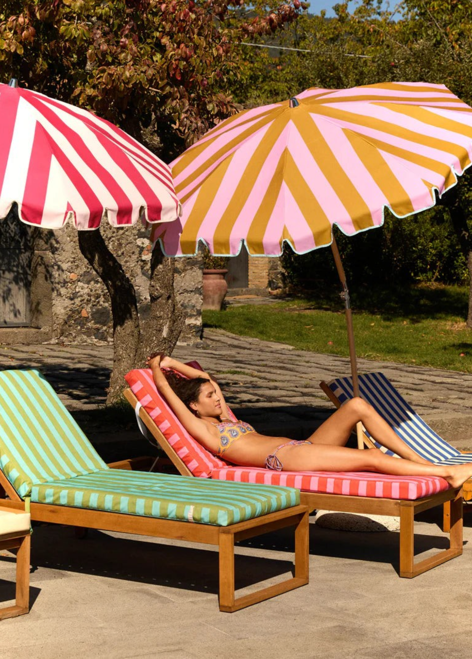 KABE SHOP INC Pietro Lizzy tie-side bikini bottom worn by a woman lounging on a striped sunbed under umbrellas.