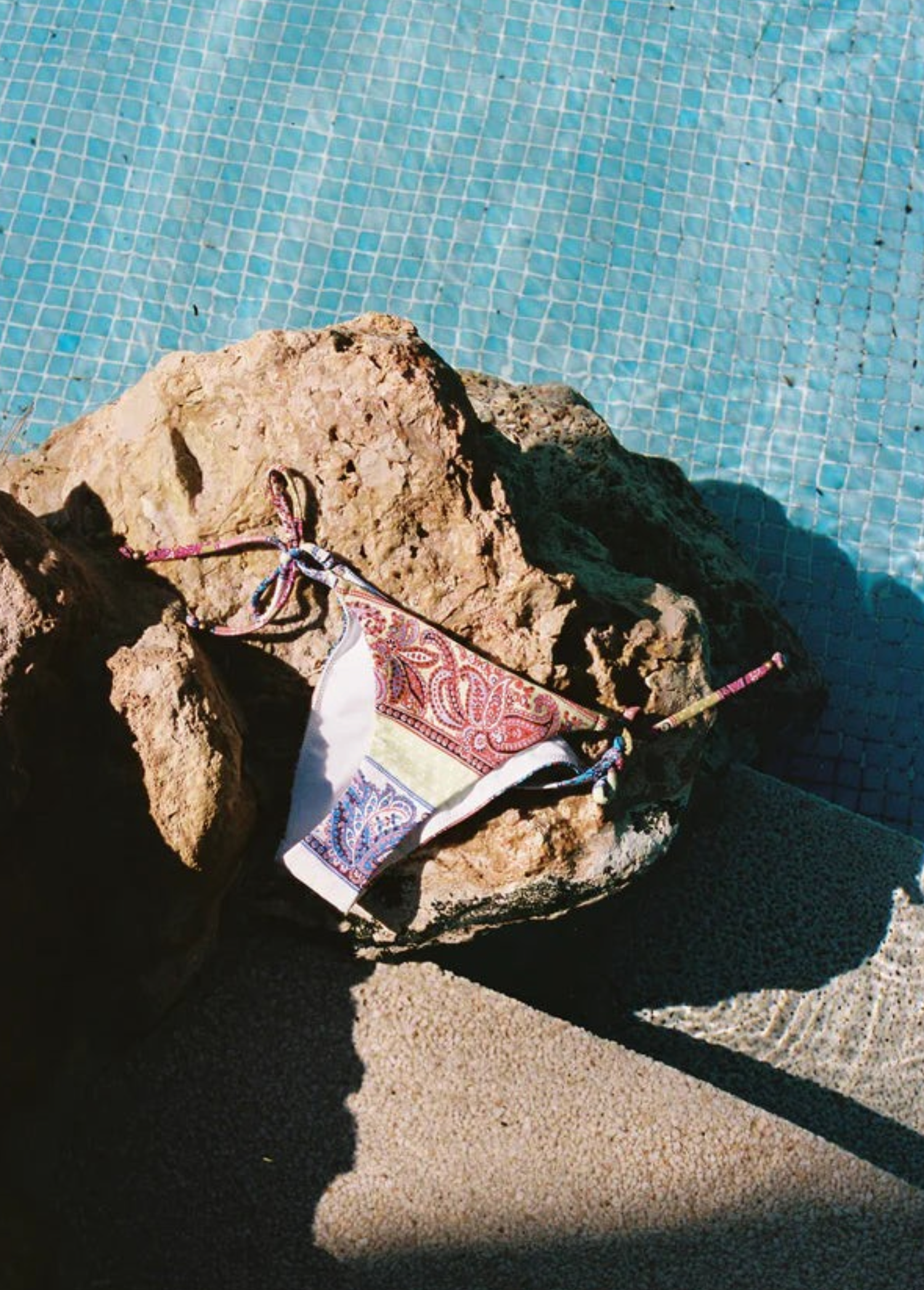 KABE SHOP INC Marseille paisley print tie-side bikini bottom resting on a rock by a blue swimming pool