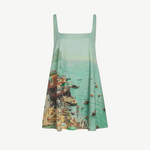 The Brio Dress is a square-neck mini dress featuring a print of a crowded beach and coastal town, designed as a versatile warm-weather essential.