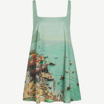 The Brio Dress is a square-neck mini dress featuring a print of a crowded beach and coastal town, designed as a versatile warm-weather essential.
