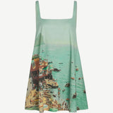 The Brio Dress is a square-neck mini dress featuring a print of a crowded beach and coastal town, designed as a versatile warm-weather essential.