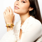 A woman with long brown hair, dressed in a white ruffled blouse, poses with her hand near her face while showcasing the Theodora Cuff—a unique gold bracelet adorned with colorful gemstones.