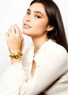 A woman with long brown hair, dressed in a white ruffled blouse, poses with her hand near her face while showcasing the Theodora Cuff—a unique gold bracelet adorned with colorful gemstones.
