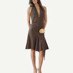 A woman wearing the Halterneck Midi Shell Dress—a sleeveless, deep V-neck brown recycled spandex dress featuring an asymmetrical hem and a tan circular waist accent—stands in open-toe heels against a white background.