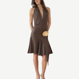 A woman wearing the Halterneck Midi Shell Dress—a sleeveless, deep V-neck brown recycled spandex dress featuring an asymmetrical hem and a tan circular waist accent—stands in open-toe heels against a white background.